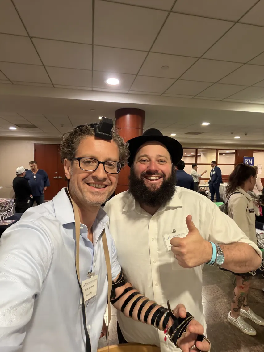 Jordan Isenstadt, wearing tefillin, takes a selfie with a smiling guest in a black hat giving a thumbs up